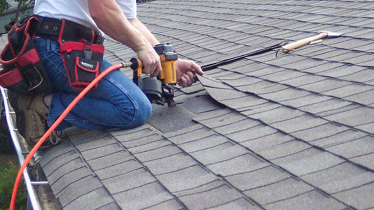 Replacing storm damaged shingles on a roof in Pittsburgh Pennsylvania using a yellow nail gun, asphalt shingles, a man in jeans and a white t shirt on the edge of the roof with his toes next to the gutters