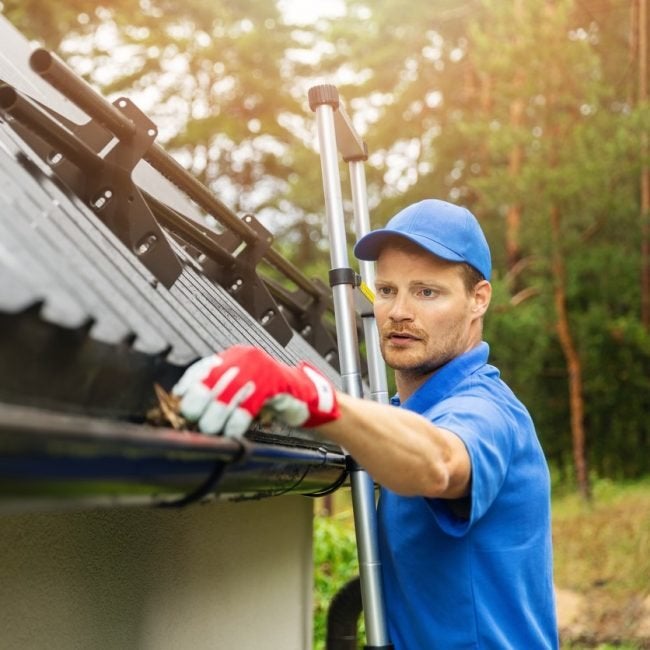 A Pit Pro Handyman in light blue shirt and cap on a ladder cleaning out a copper gutter using gloves. 