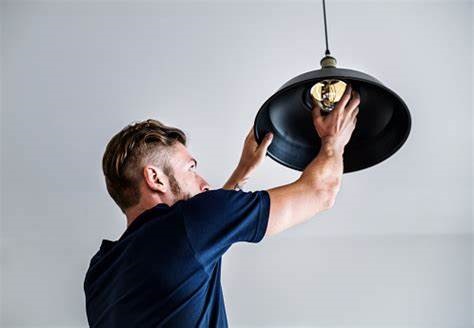 A Pit Pro Handyman on a ladder, in a blue shirt, changing a light bulb in an old fashion style warehouse dangling light.