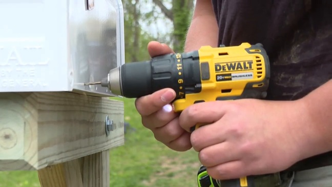 A Pit Pro Handyman installs a mailbox on a wood mail box post using a DeWalt drill. The image is a close up with a metal mailbox that is rural style.