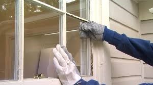 A Pit Pro Handyman replacing a window pane that was broken by a flying rock that was thrown by a lawn mower. Bjue arms and gloves mans arms are placing the glass pane in the broken pane area. 