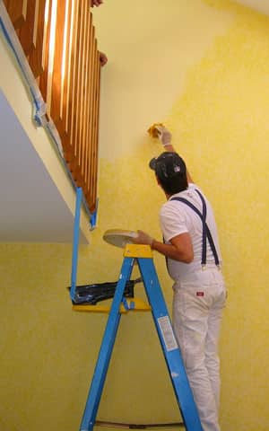 Interior Painting Handyman in Pittsburgh PA painting a wall yellow, on a ladder with a wood balcony above that is not painted. 