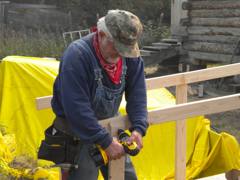 A Pit Pro Handyman using a drill and screws to construct a handicap accessible wood ramp at a historic log house. The ramp features ADA compliant rails and slope. A yellow tarp is in the background covering the supplies. 