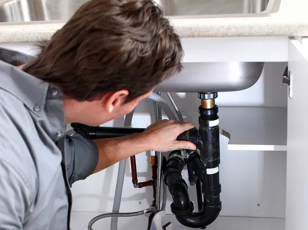 A Pit Professional Handyman inspecting the under-sink pipe for a leak on a client's leaky commercial kitchen plumbing repair. A silver sink bottom and white cabinet and metal counter top