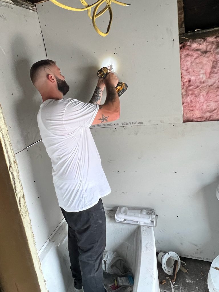 Pit Pro Handyman hanging sheet drywall in a bathroom remodel project located in Cranberry Township. A Handyman with drill in his hands screwing sheet of drywall to wood studs. 
